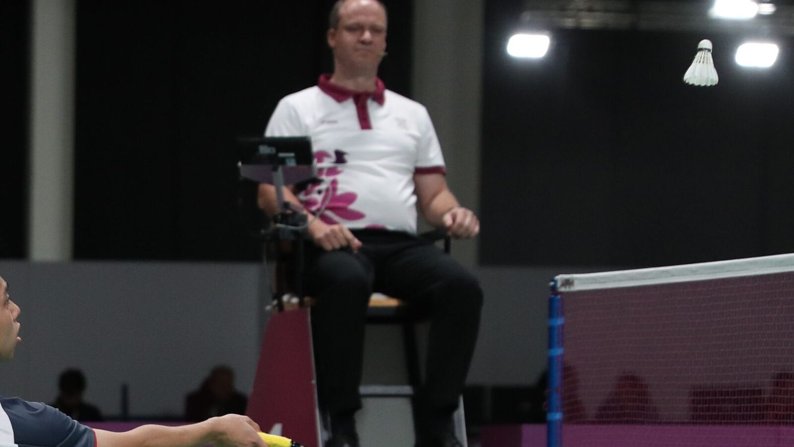 Une personne arbitre de badminton est assise sur la chaise surélevée près du filet et observe un échange pendant que le volant passe devant elle.