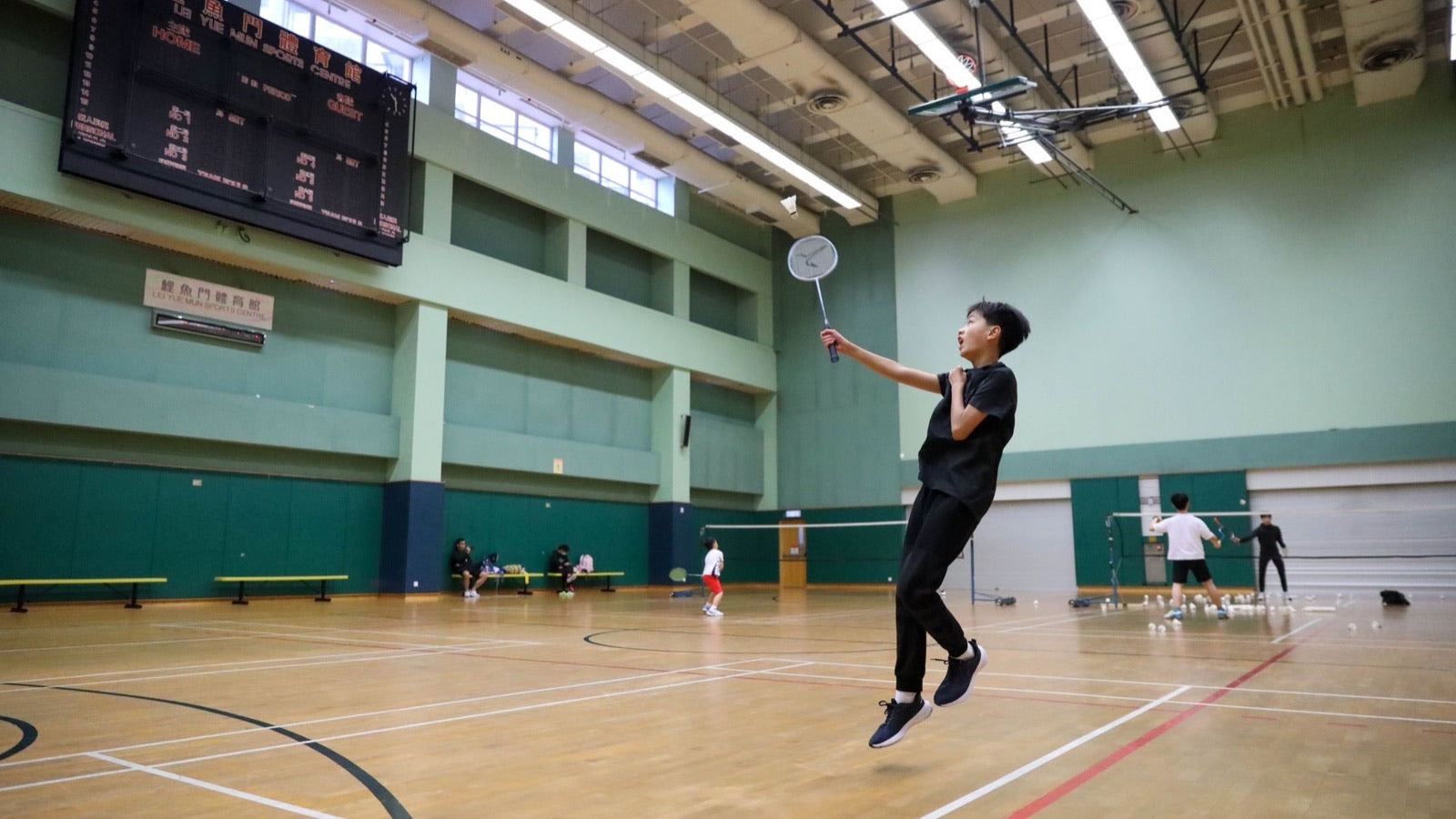 Une jeune joueuse de badminton s’élève haut au-dessus du terrain pour smasher, la raquette levée au-dessus de la tête, tandis que d’autres personnes s’entraînent à l’arrière-plan d’un centre sportif intérieur.