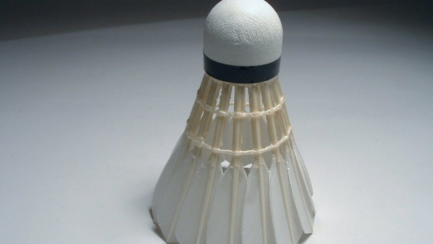 Un seul volant blanc en plumes, avec une base en liège cerclée de noir, posé à la verticale sur un fond gris uni de studio.