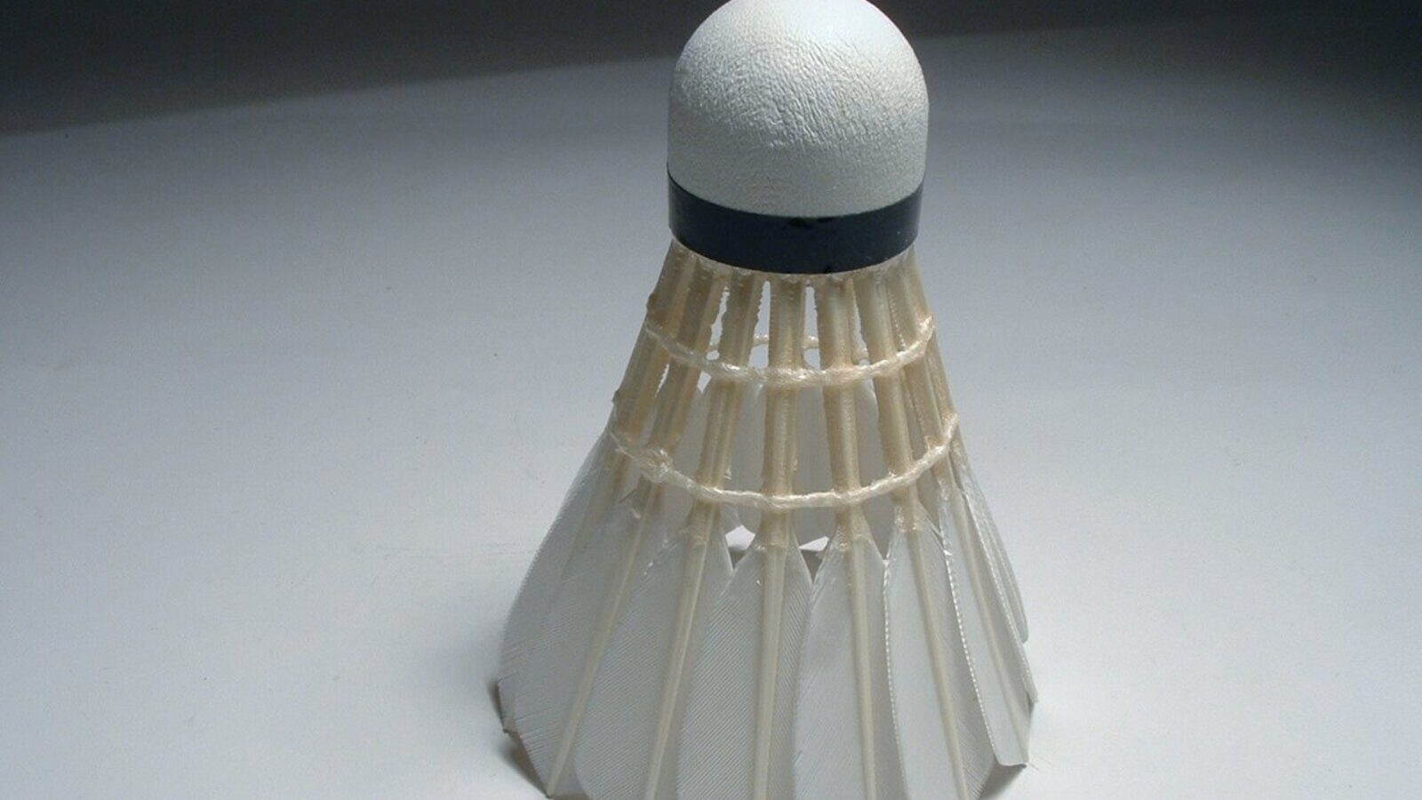 Un seul volant blanc en plumes, avec une base en liège cerclée de noir, posé à la verticale sur un fond gris uni de studio.