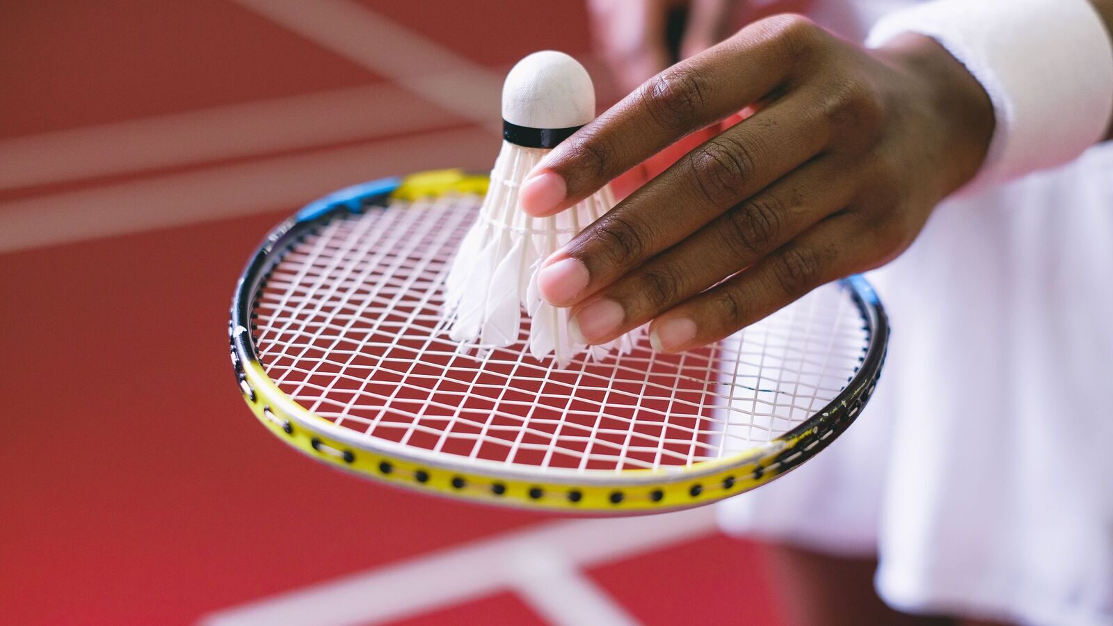 Gros plan sur le cordage et le pourtour d’une raquette de badminton pendant qu’une main pose un volant sur les cordes; terrain intérieur en arrière-plan.