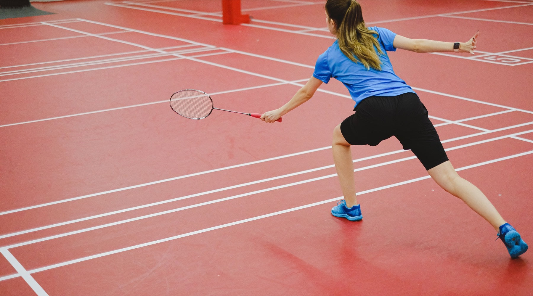 Personne jouant en simple, vue de dos, qui fait une fente pour frapper un coup droit sur un terrain de badminton intérieur.