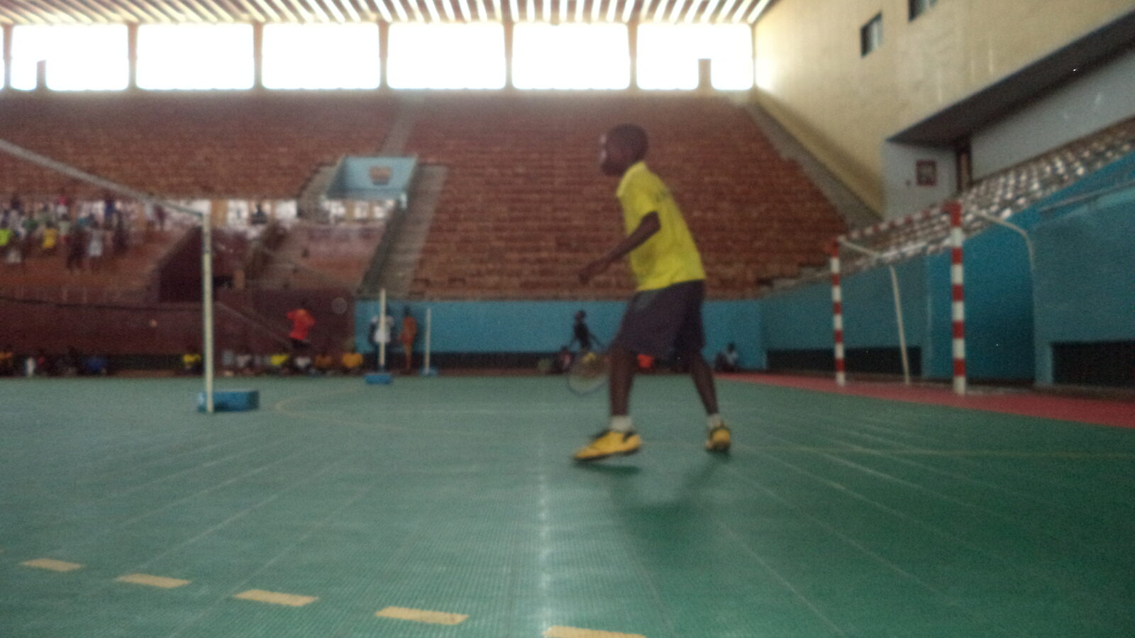 Une joueuse ou un joueur de badminton en chandail jaune se déplace sur le plancher vert d’un gymnase polyvalent, raquette à la main, avec des gradins vides et des buts de handball en arrière-plan.