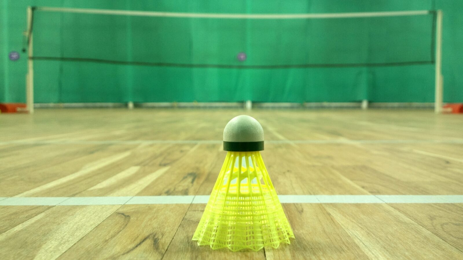 Un volant jaune repose sur un terrain de badminton intérieur en bois, le filet et les poteaux visibles en arrière-plan, flous.