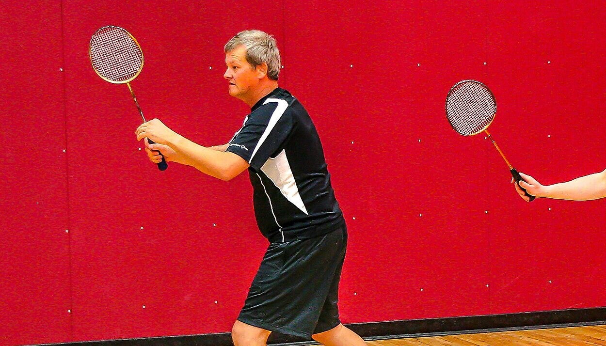 Deux personnes qui jouent au badminton dans un gymnase intérieur tiennent leur raquette en position d’attente entre les points, devant un mur matelassé rouge et un plancher de bois.