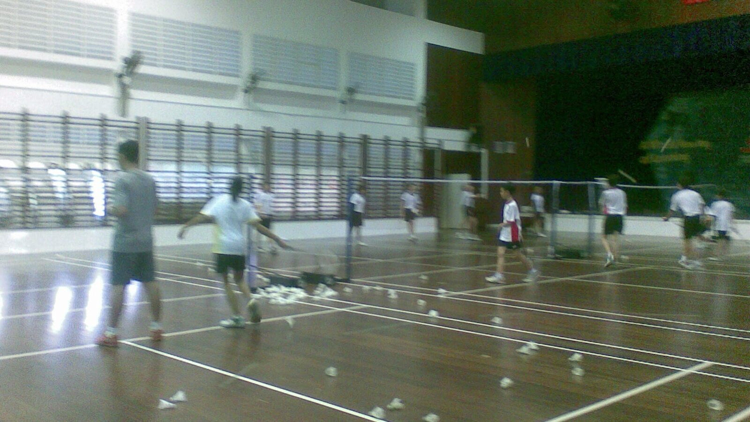 Des élèves jouent au badminton sur plusieurs terrains dans un gymnase scolaire, avec des volants usagés éparpillés au sol après un exercice de services.