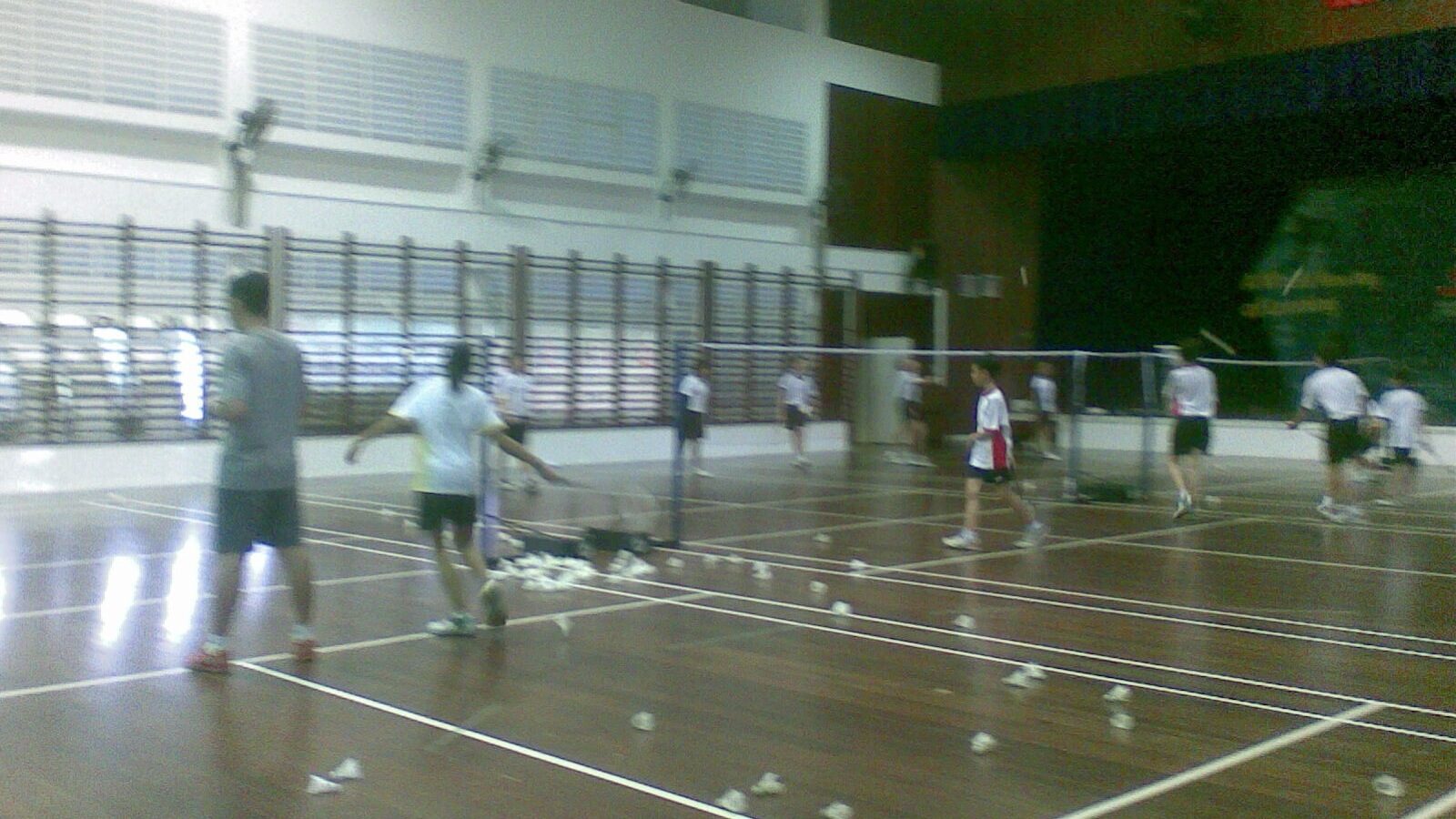Des élèves jouent au badminton sur plusieurs terrains dans un gymnase scolaire, avec des volants usagés éparpillés au sol après un exercice de services.